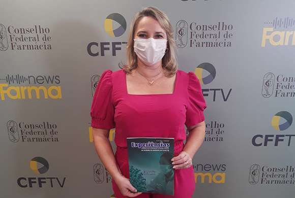 Dra. Luciana Canetto, segura um exemplar da revista, veste uma roupa rosa e usa máscara. Ao fundo um totem com logotipos do CFF