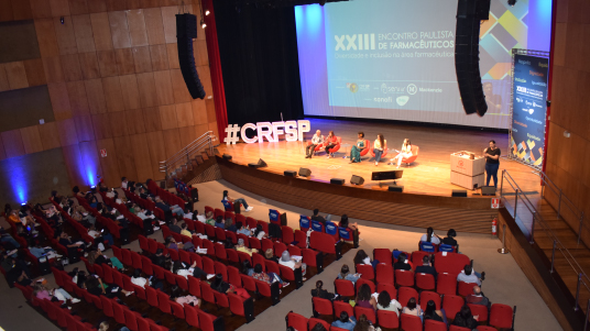 Imagem do alto com auditório com cadeiras vermelhas e pessoas sentadas em frente ao palco com as cadeiras para palestrantes, um telão e um logotipo do CRF-SP iluminado