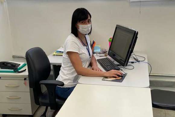 Dra. Margareth Ferreira Cunha sentada em frente a um computador sobre uma mesa branca e usando máscara
