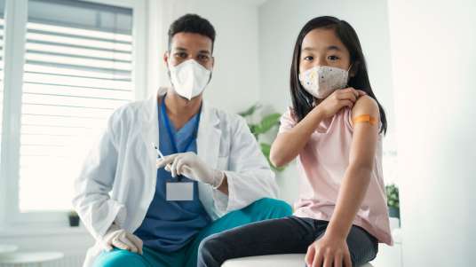 Farmacêutico jovem de mascara segura uma seringa de vacina, ao seu lado uma criança oriental de máscara está com a manga da camiseta levantada e um curativo no braço, indicando que foi vacinada