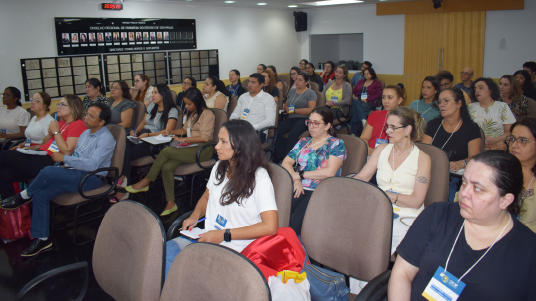 Foto de auditório com público diversificado, composto com pessoas de etnias e faixas etárias diferentes