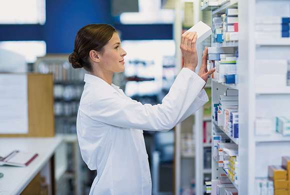 Foto de mulher branca paramentada de farmacêutica, mexendo nos estoques de uma farmácia.