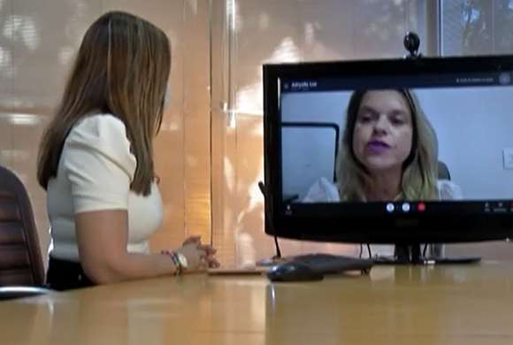 Uma mulher de lado com uma blusa branca sentada em uma cadeira em uma mesa de madeira entrevistando uma mulher loira que está em um monitor de computador