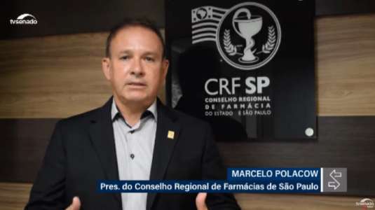 Dr. Marcelo Polacow (branco, cabelo curto, blazer preto e camisa social cinza) em pé em frente ao logo do CRF-SP que está na parede 