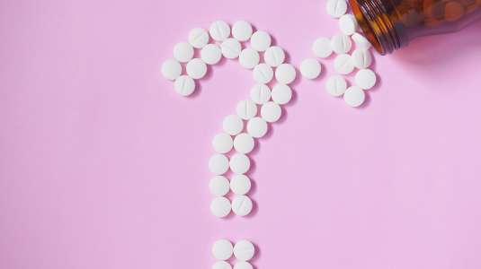 Um fundo rosa com um frasco de medicamento aberto caído e saindo comprimidos brancos formando um ponto de interrogação