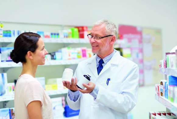 Farmacêutico grisalho e de óculos de jaleco branco conversa com mulher de camise ta clara e cabelo preso, ambos estão em pé dentro da farmácia  