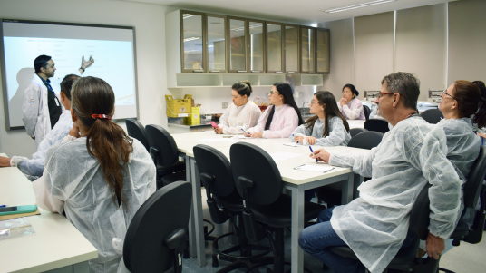 Alunos vestidos com jaleco branco sentados em cadeiras prestam atenção em aula de Dr. Claudinei, que está à frente apontando para um quadro branco