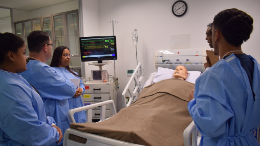 Foto com um boneco realístico de um paciente na uti, deitado em uma cama hospitalar, com um cobertor marrom em cima. quatro pessoas de lado com aventais azuis