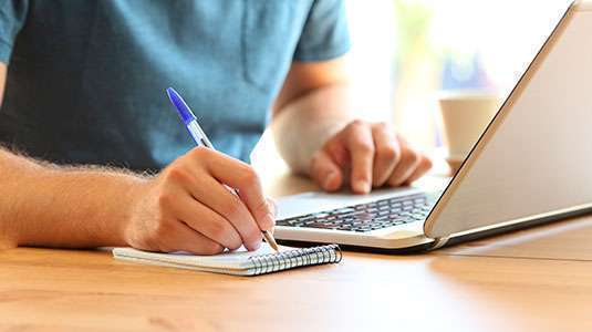Homem escrevendo em caderno ao lado de laptop
