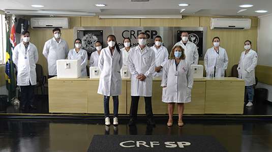 Doze pessoas com jaleco entre homens e mulheres de pé na frente de uma parede com o logo do CRF-SP