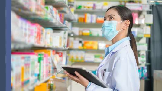Farmacêutica branca de jaleco e máscara, cabelos presos, está com uma prancheta na mão olhando para os medicamentos na prateleira de uma farmácia 