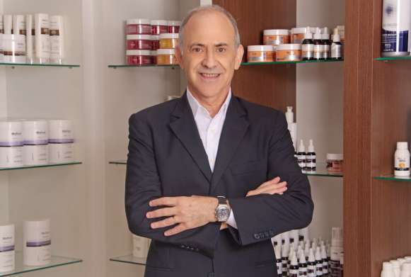 Dr. Marcelo Schulman é um homem branco, calvo, de blazer preto e camisa branca, de braços cruzados e à frente de uma prateleira de produtos da Vitaderm