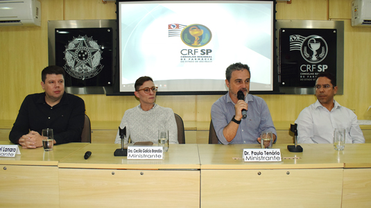 Foto de mesa de debates feita de madeira clara, tendo ao fundo tela de projetor com o logo do CRF-SP. Sentados estão três homens e uma mulher; dois homens são brancos e um não branco. A mulher é branca